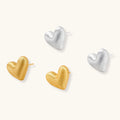 Pair of oversized brushed gold heart stud earrings with a puffy, sculpted surface and post-back closure on a cream background.