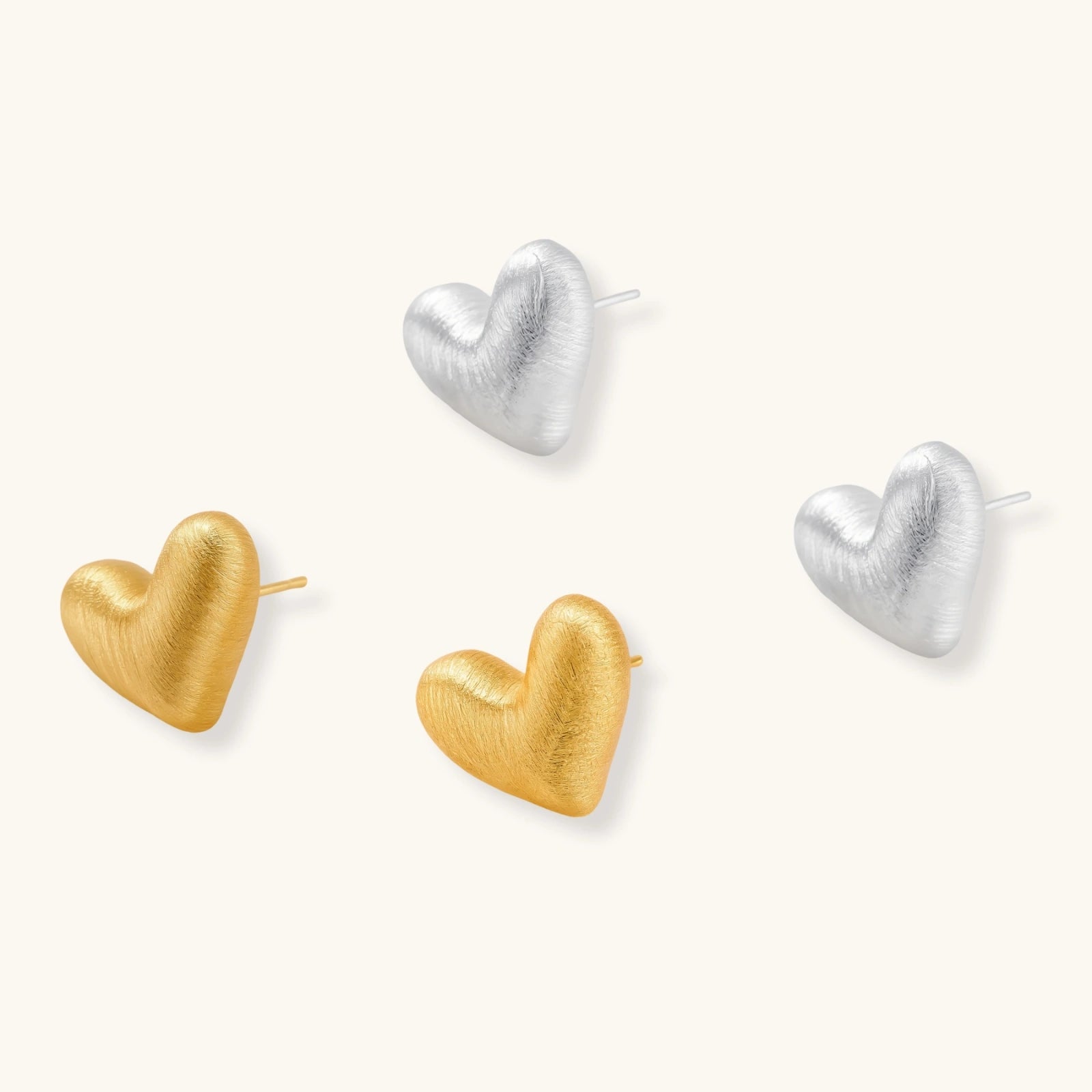 Pair of oversized brushed gold heart stud earrings with a puffy, sculpted surface and post-back closure on a cream background.